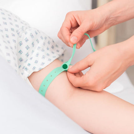 A mint green medical identification wristband being applied to a person's forearm by a healthcare provider's hands. The person is wearing a white hospital gown with a blue geometric pattern.