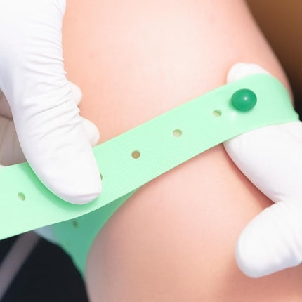 A mint green medical identification wristband with a green snap button being applied to a person's forearm by gloved hands.