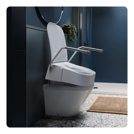 A white toilet with a raised seat and attached metal grab handles, shown installed in a modern bathroom with dark blue walls, speckled gray floor, and a small woven mat.