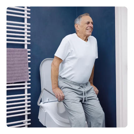 An older man in a white t-shirt and gray sweatpants sits on a white toilet in a bathroom with a dark blue wall and a white towel radiator.